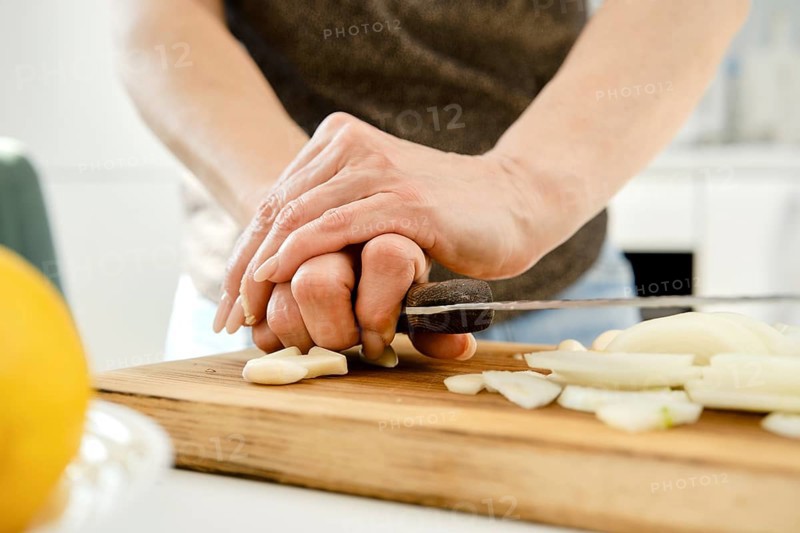Unrecognizable woman crush garlic to finely chop it for sauce on a wooden cutting board in a bright kitchen