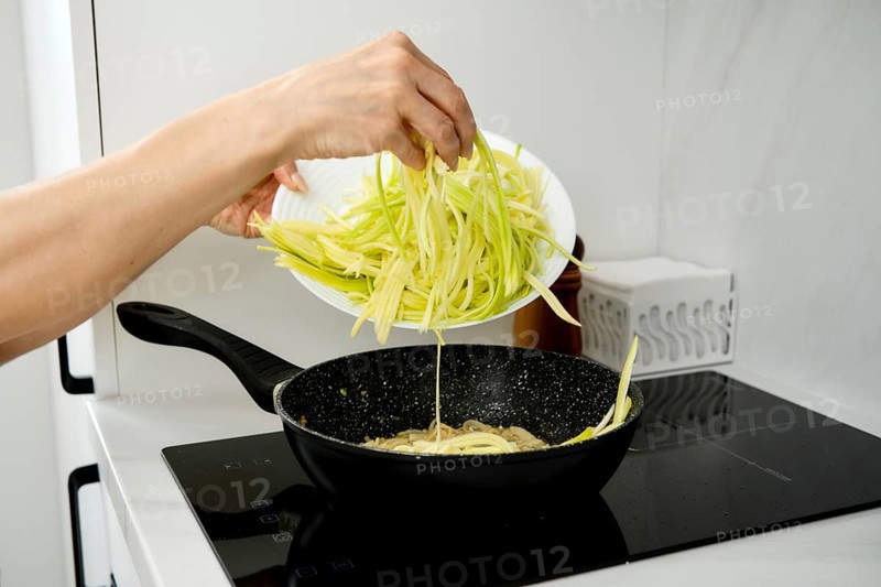 A hand pours freshly cut zucchini noodles into a sizzling pan on an electric stovetop. The kitchen is bright and modern, showcasing meal preparation for an evening meal
