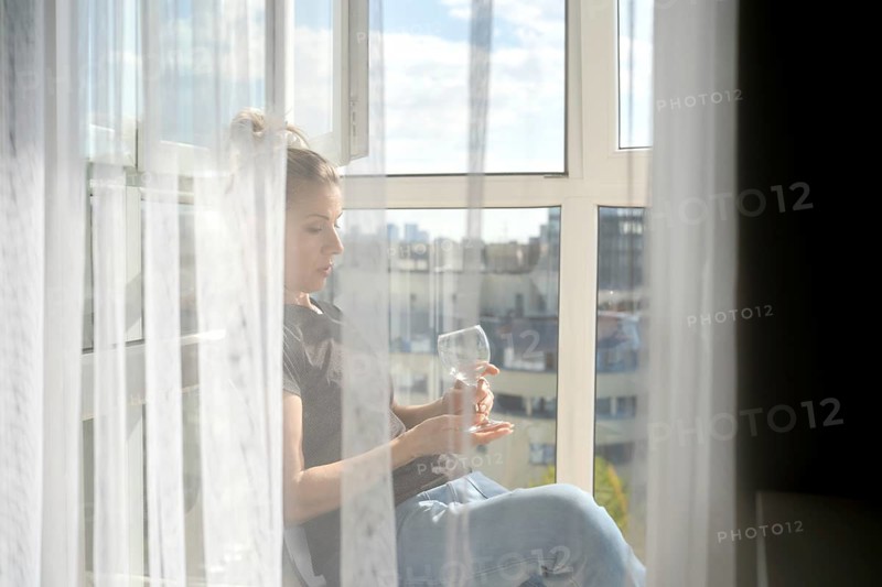 Sunlight streams through sheer curtains as a woman sits peacefully by the window. She gazes thoughtfully at a glass, enjoying the tranquil moment while overlooking a bustling city