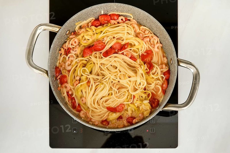 A pot of spaghetti is simmering on a stove, mixed with shrimp and cherry tomatoes