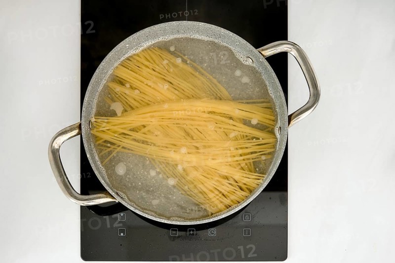Long strands of pasta are submerged in boiling water inside a pot on an induction stovetop. The kitchen environment is clean and modern, showcasing the cooking process clearly
