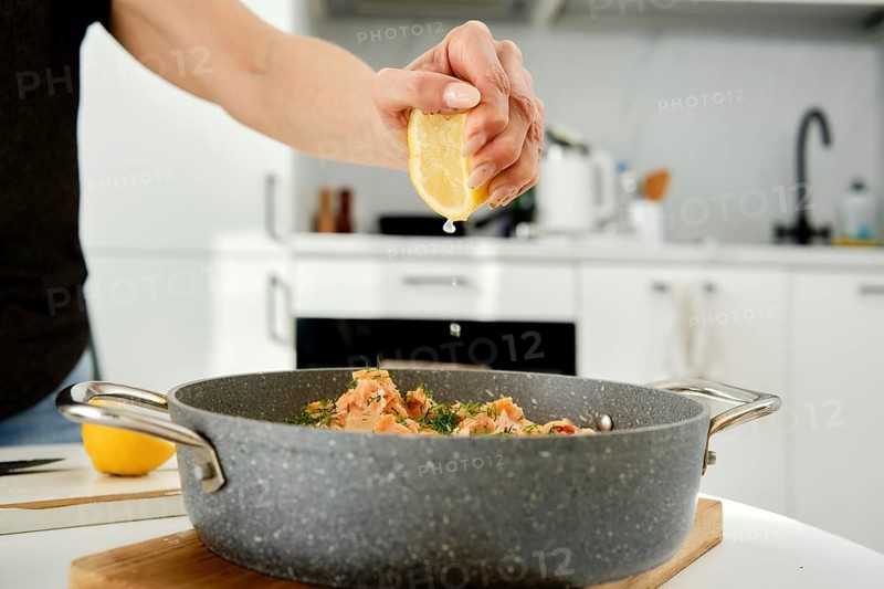 A person is preparing a seafood dish by squeezing fresh lemon juice into a skillet. The modern kitchen features sleek appliances and a vibrant atmosphere, enhancing the cooking experience