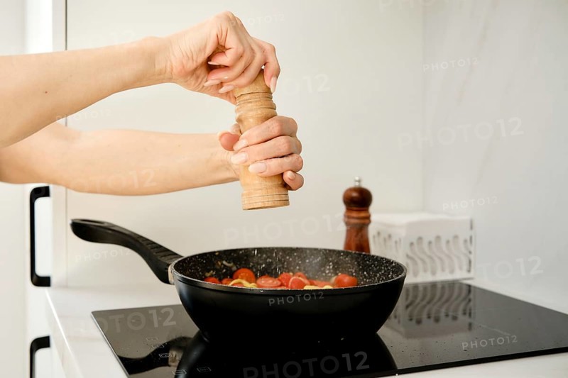 A hand is grinding pepper over a skillet filled with fresh tomatoes. The setting is a modern kitchen featuring a sleek countertop and cooking utensils, creating a warm cooking atmosphere