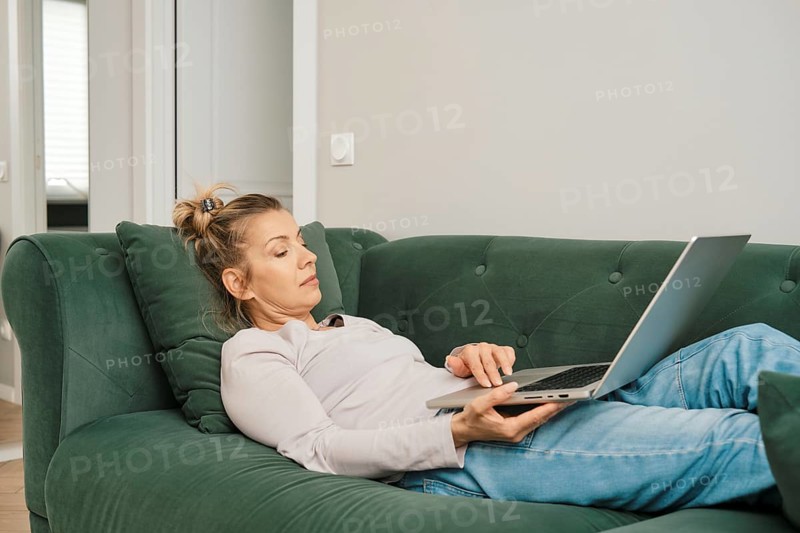A woman with a relaxed demeanor is laying on a green couch in a bright living room, focused on her laptop