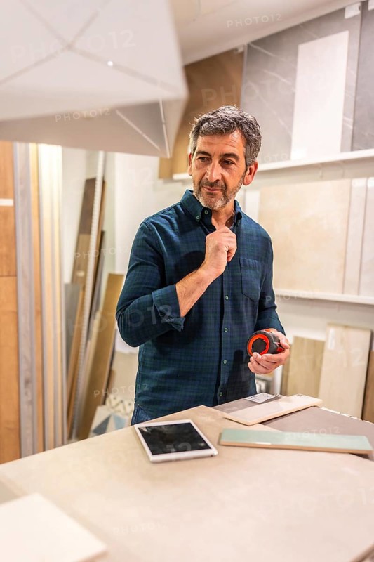 Mature architect holding measuring tape and choosing ceramic tiles and wooden panels for interior design project in home improvement store