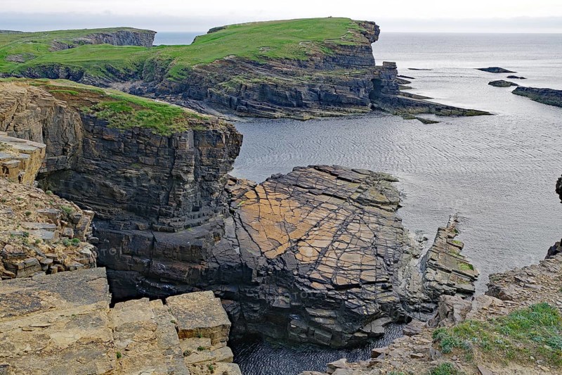 Dramatic rocky coastline with green cliffs and calm sea under a cloudy sky, Orkney Islands, Scotland, United Kingdom