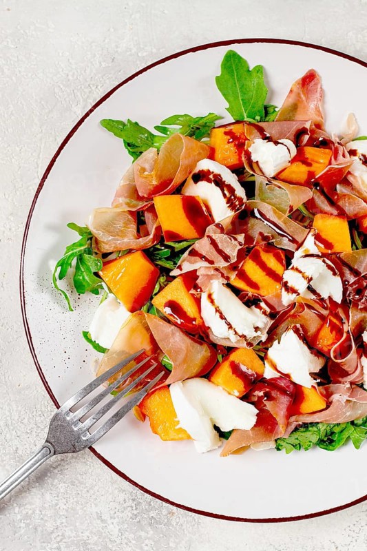 Salad with slices of ripe melon, prosciutto, mozzarella and fresh arugula, decorated with balsamic glaze