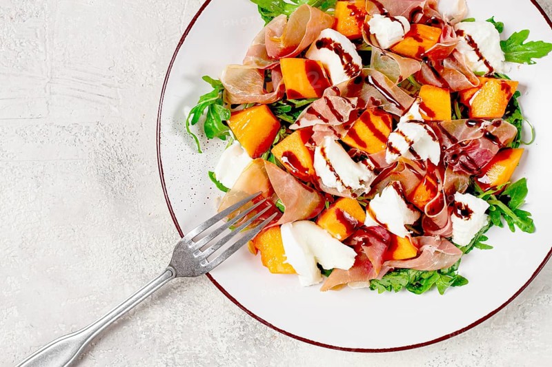 Salad with slices of ripe melon, prosciutto, mozzarella and fresh arugula, decorated with balsamic glaze