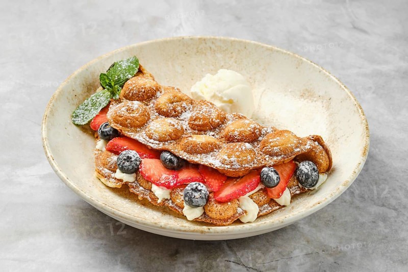 Colorful bubble waffles are served with fresh strawberries and blueberries, topped with creamy filling and a scoop of ice cream. The dish is garnished with mint leaves for a refreshing touch