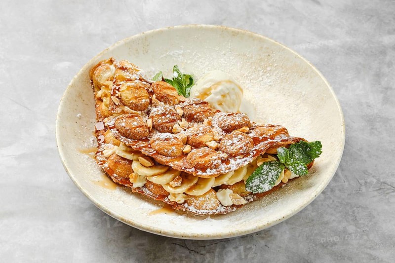 A beautifully presented dessert showcases a crispy waffle filled with sliced bananas and caramel, topped with powdered sugar and a scoop of vanilla ice cream. Mint leaves add a pop of color