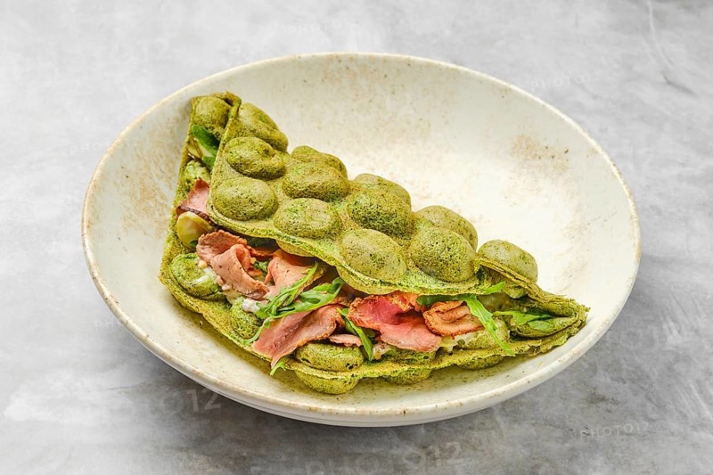 A matcha pancake showcases a vibrant green hue, folded elegantly with layers of savory ingredients such as slices of meat and fresh greens