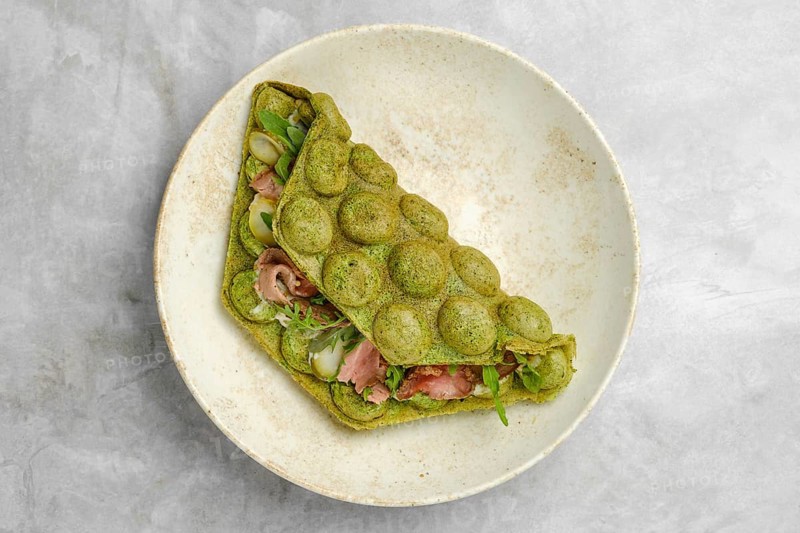 A green wrap made with spinach or herbs contains layers of fresh vegetables and meat, resting on a simple plate. This dish is presented in a relaxed cafe setting, perfect for a light meal
