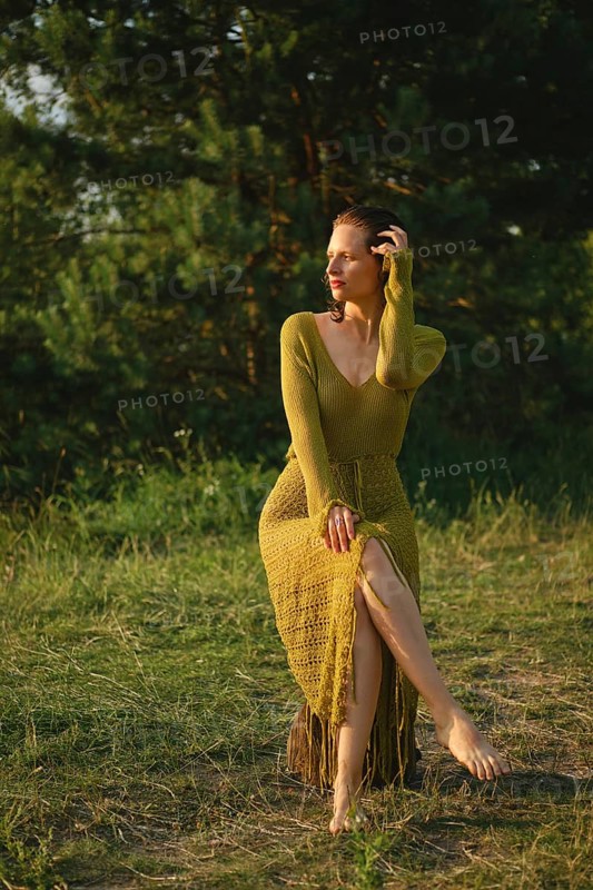 On a warm day, a woman sits gracefully by the lake surrounded by lush greenery. Wearing a fishnet knitted dress, she enjoys the tranquility of nature and the refreshing atmosphere of summer