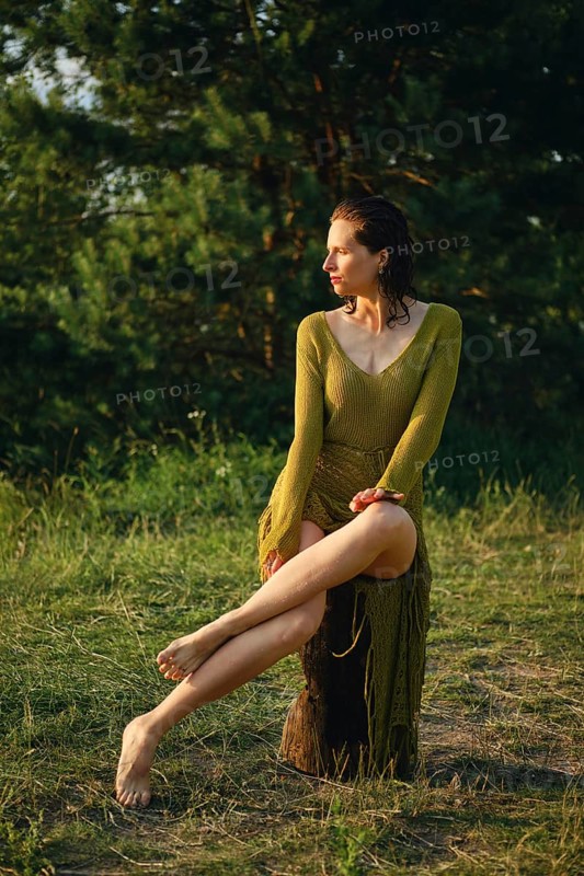 A woman enjoys a hot day by the lake, sitting gracefully on a rustic log. The sun casts a warm glow on her, highlighting her relaxed pose amidst lush greenery