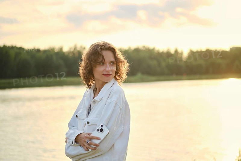 Cute woman with curly hair stands by the lake at sunset. She look back while holding smartphone in her hand