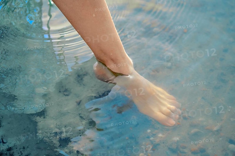 One foot dips into the refreshing lake water, creating gentle ripples on the surface. The bright sun shines down, making the day perfect for enjoyable outdoor activities and relaxation by the water