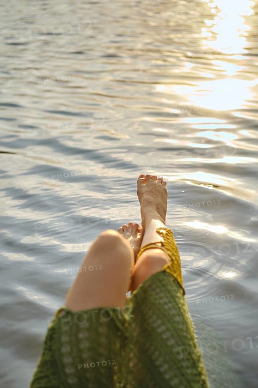 Enjoying a hot day at the lake, woman relaxes with her feet dipped in the calming water. The sun creates sparkling reflections, enhancing the tranquil atmosphere of the moment