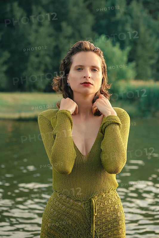 Bright sunlight casts a warm glow on a serene lake, where a woman poses gracefully in a green outfit. Lush greenery reflects in the water, creating a peaceful atmosphere on this hot day