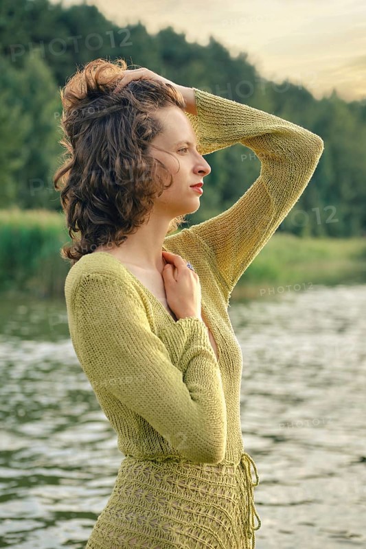 On a hot day, a woman stands gracefully by a serene lake touching her curly hair. She seems lost in thought while embracing the beauty of her natural surroundings