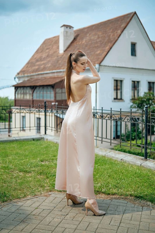 A young woman showcases her stylish outfit in a soft-colored dress while standing in a grassy area. Behind her is a charming vintage house with classic architecture, all under a bright sky