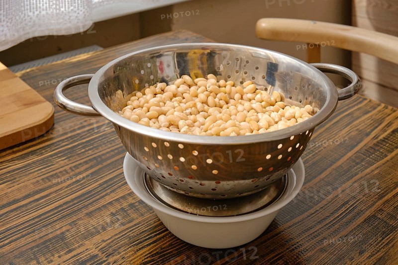 A colander filled with soaked white beans is resting above a bowl to catch excess water. Natural light filters into the cozy kitchen, highlighting the rustic wooden surface and homey atmosphere