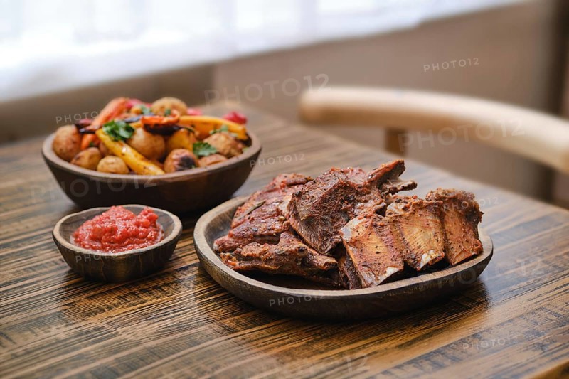 Tender roasted short beef ribs on a wooden table alongside a bowl of colorful roasted vegetables and a small dish of salsa. A cozy indoor atmosphere enhances the meal's appeal