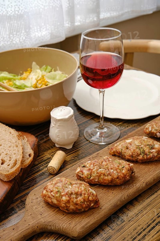 A table is set with a wooden board of homemade patties, a bowl of fresh salad, a glass of red wine, and slices of bread. Sunlight streams through the window, enhancing the cozy atmosphere