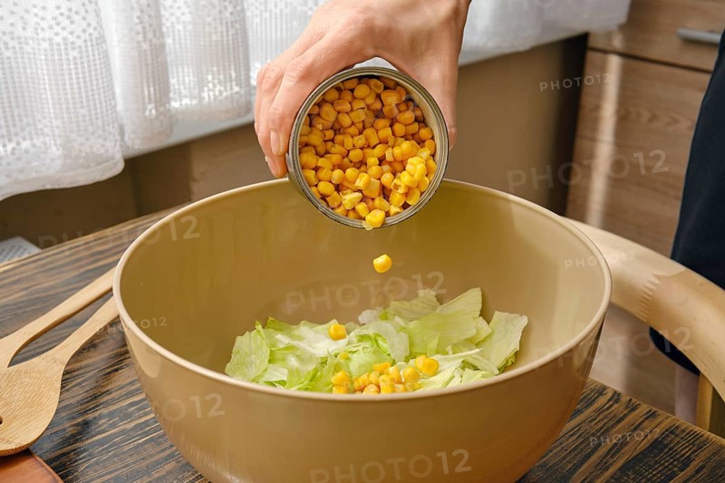 A hand pours canned corn into a bowl filled with fresh green lettuce. The warm kitchen features wood accents, creating a welcoming atmosphere for meal preparation