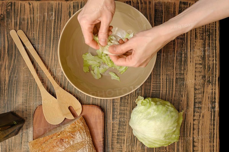 Hands tear crisp lettuce leaves into a bowl, surrounded by wooden utensils and fresh ingredients. The rustic table adds warmth to this vibrant and healthy preparation activity