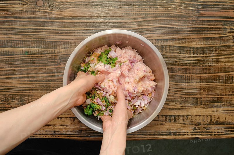 Hands are mixing raw minced meat, chopped onions, herbs, and spices in a stainless steel bowl on a rustic wooden countertop. The preparation indicates a focus on creating a traditional cutlet