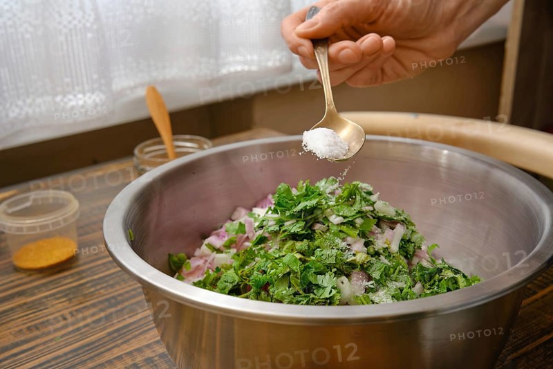 A hand adds salt to a bowl filled with minced meat, chopped onions and fresh herbs. The kitchen is bright, with sunlight streaming through the window, creating a warm atmosphere for cooking