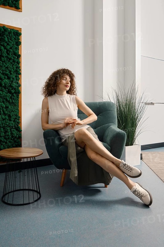 Woman relaxing in a stylish chair in a modern office space with greenery and natural light. She has curly hair and wears a casual outfit suitable for everyday wear