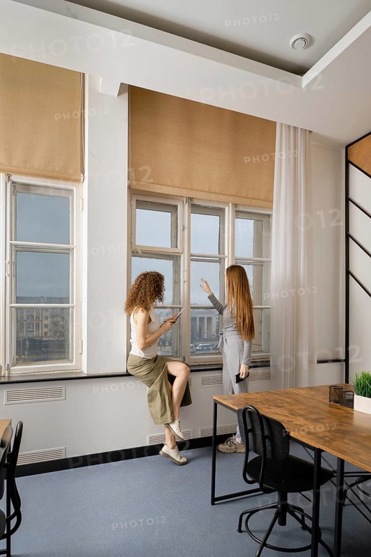 Two women engage in a conversation while standing by large windows in a modern office setting. The atmosphere is professional and vibrant, emphasizing collaboration in women's health initiatives
