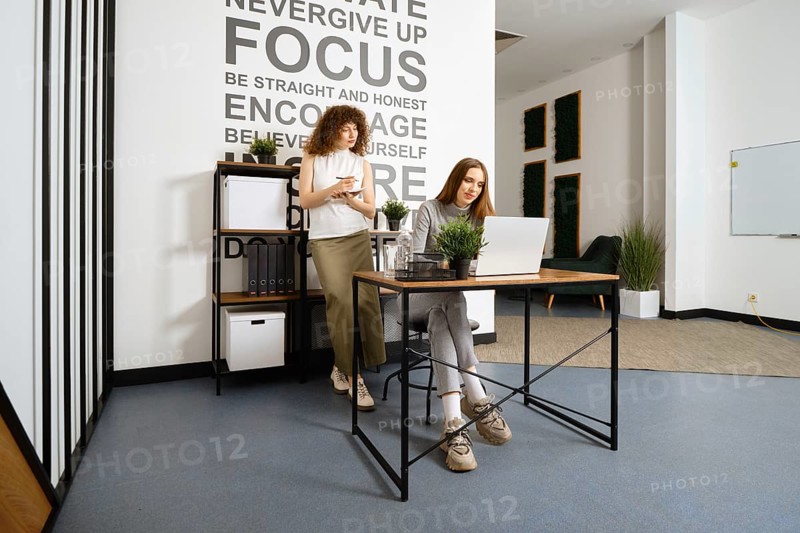 Two women working in bright office. One takes notes while the other types on a laptop, showcasing teamwork and innovative ideas