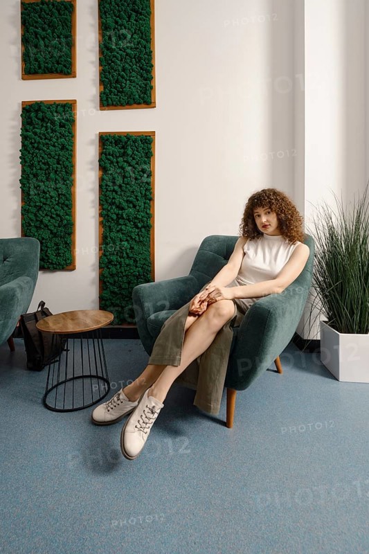 A young woman sits comfortably in a contemporary chair, dressed in a casual outfit. The room features greenery on the walls, stylish furniture, and minimalist decor, creating a serene atmosphere