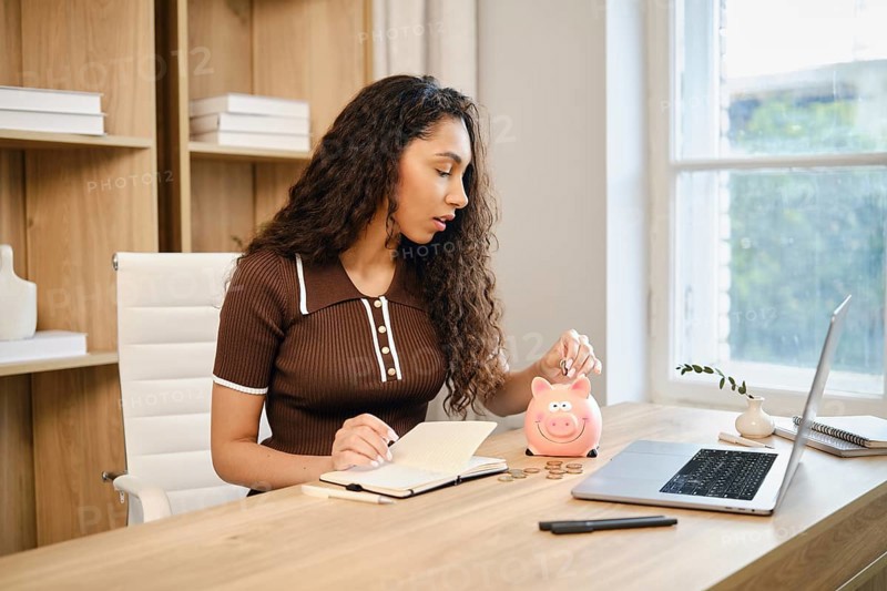 A woman sits at a wooden desk in a stylish home office, writing in a notebook while dropping coins into a pink piggy bank. Natural light filters through the window, creating a warm atmosphere