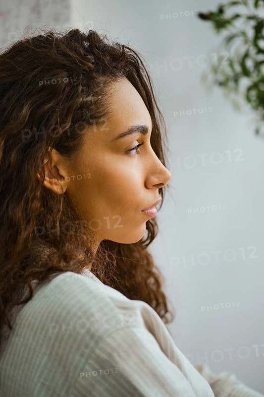 In a cozy indoor space, a woman with long, curly hair sits in profile, lost in thought. Soft natural light highlights her features, creating a serene atmosphere filled with tranquility