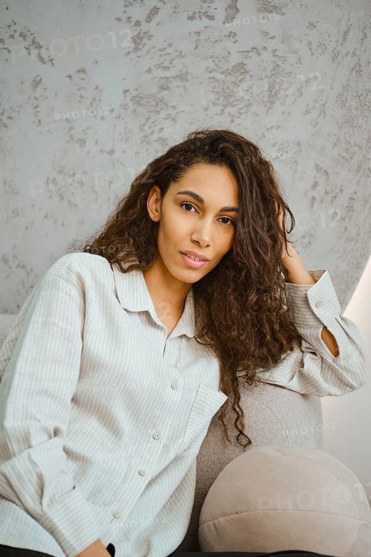 Charming woman with curly hair sits comfortably on a plush sofa in a contemporary indoor space. Sunlight filters through the window, highlighting her relaxed pose and fashionable attire