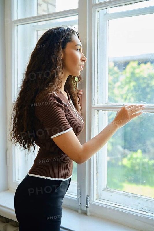 A woman admires the greenery outside while standing by a window. Her curly hair cascades down her shoulders, and she appears contemplative in the warm afternoon light