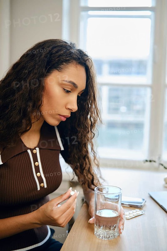 A young woman with curly hair sits at a table concentrating on a pill in her hand and a glass of water beside her. Sunlight filters through the window, creating a serene atmosphere