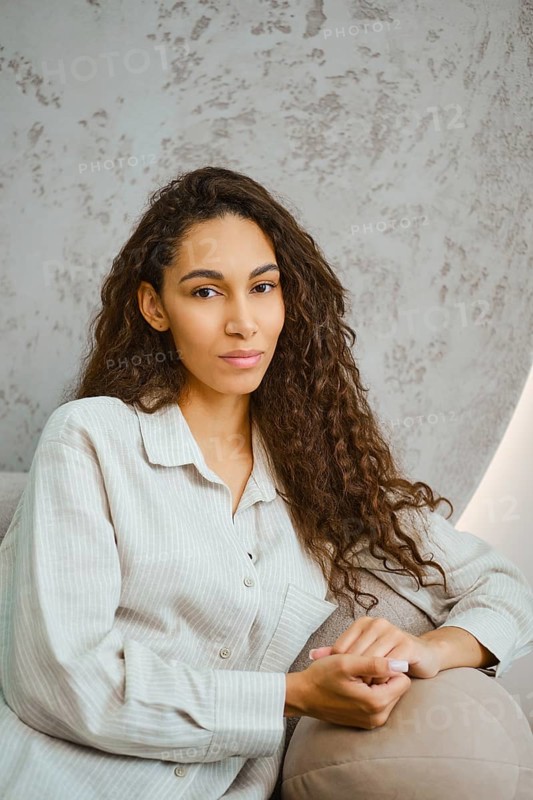 A young woman with long, curly hair is lounging on a sofa in a contemporary space. Soft afternoon light fills the room, highlighting her thoughtful expression and the elegant design around her
