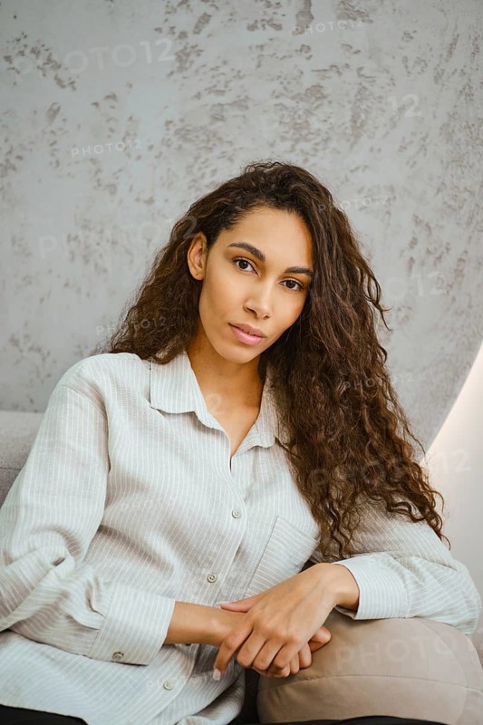 A young woman with long curly hair poses casually in a softly lit modern interior. She rests her arm on armrest of a sofa, exuding a sense of confidence and comfort, dressed in a stylish button-up shirt