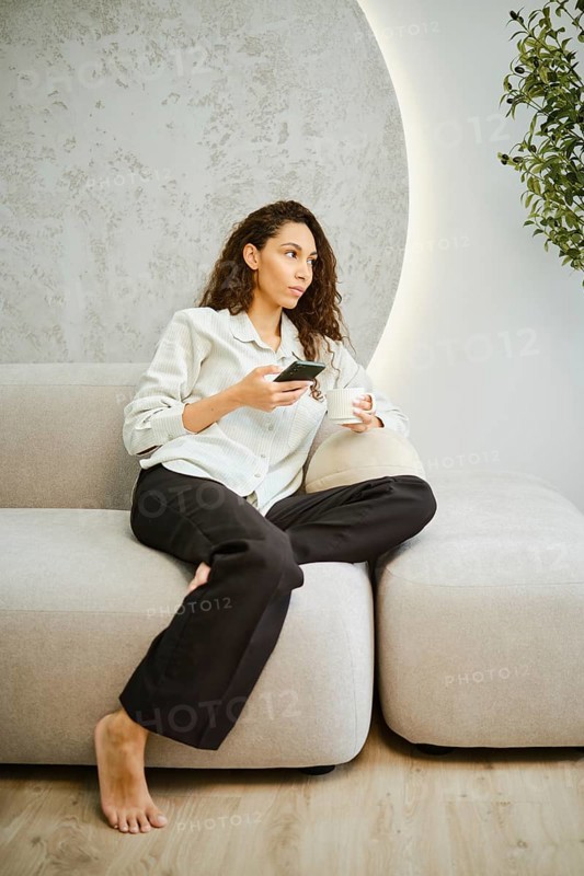 A woman with curly hair sits comfortably on a light sofa in a stylish living room, holding a phone and deep in thought. The soft lighting and minimalist decor enhance the cozy atmosphere