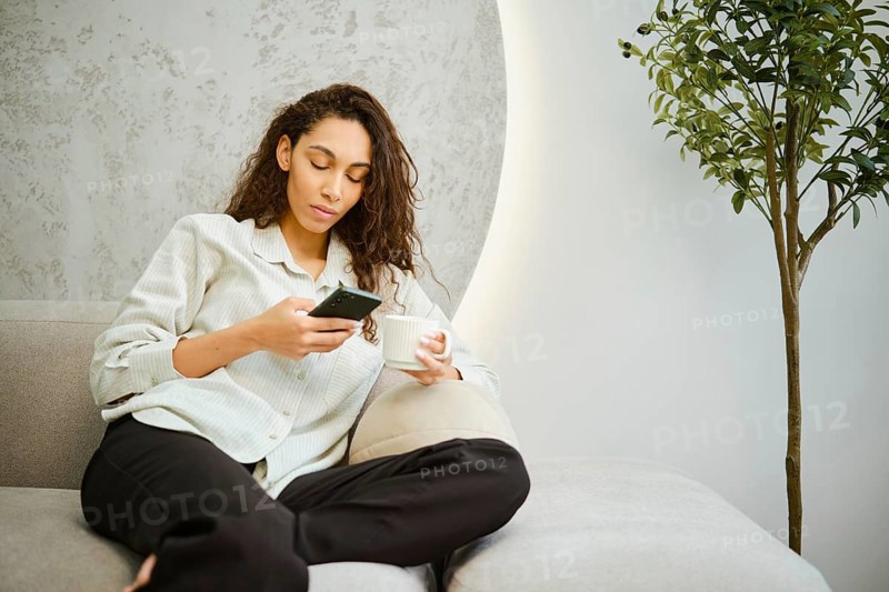 A young woman sits comfortably on a couch in a modern living room, focused on her smartphone while holding a cup. Natural light enhances the relaxed atmosphere of the setting