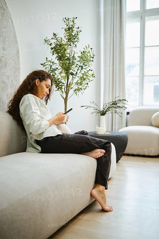 A barefoot woman sits comfortably on a beige sofa, engrossed in her smartphone. The bright living room features natural light, plants, and minimalist decor that enhances the inviting atmosphere