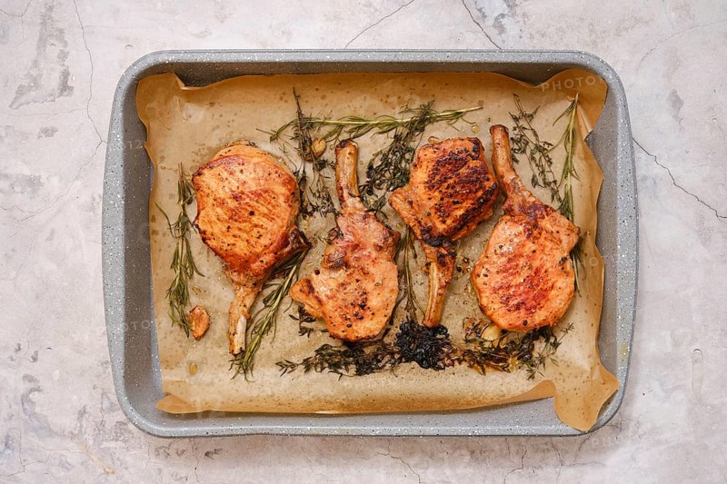 Juicy pork rib roasts are seasoned and cooked to perfection on a baking tray lined with parchment paper. Fresh herbs add flavor, creating an appetizing dish suitable for any occasion