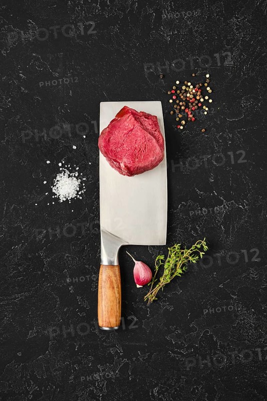 Fresh meat sits atop a large knife on a dark countertop, surrounded by salt, pepper, herbs, and garlic. The setting suggests preparation for a delicious meal, showcasing culinary artistry