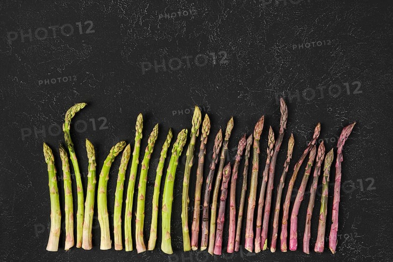Different varieties of fresh asparagus are arranged neatly on a dark textured surface. The vibrant greens and purples create a colorful contrast, showcasing the freshness and quality of the produce