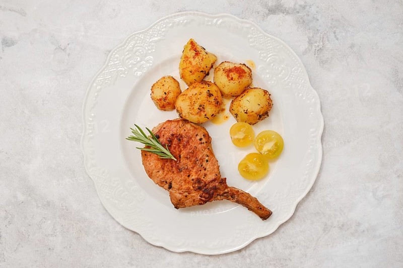 A roasted pork rib roast on a decorative white plate served with golden roasted potatoes and sweet yellow cherry tomatoes