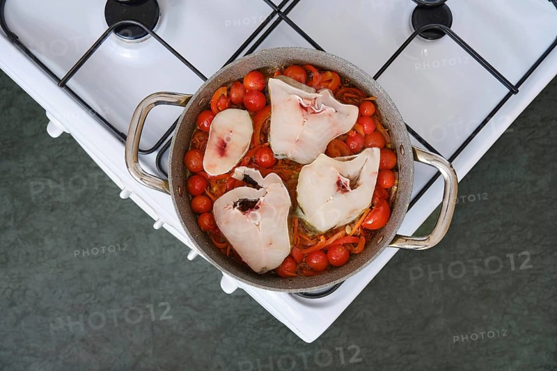 Bright red tomatoes and colorful peppers surround large pieces of cod fillet in a pot on a stove